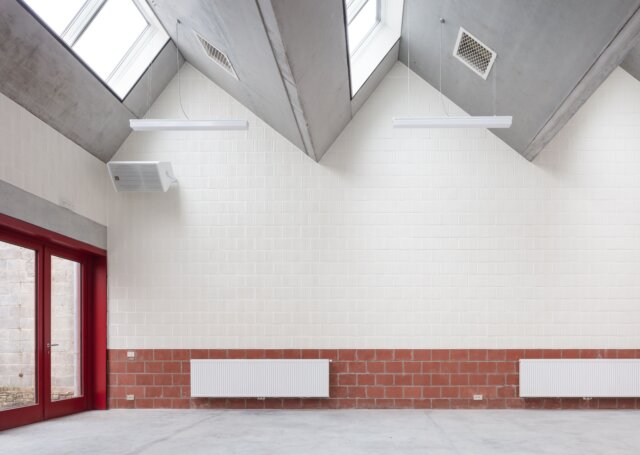 ruimte met betonnen puntdak, deur in rood schrijnwerk, witte tegelwand en bakstenen plint