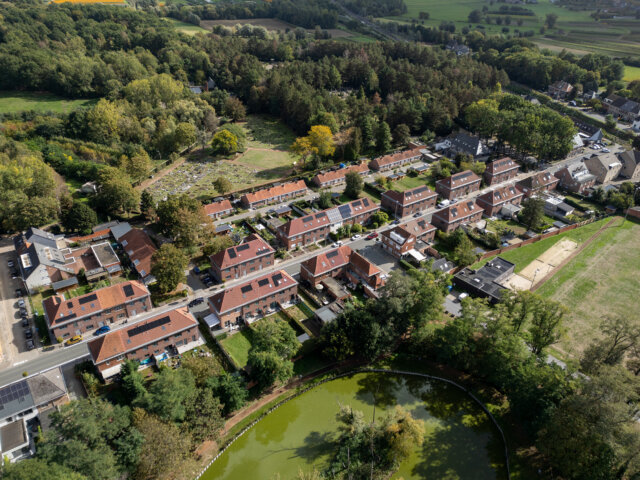 Schuine luchtfoto van de sociale woonwijk in Wetteren, met rijen huizen met rode pannendaken tussen bomen, grasvelden en een vijver