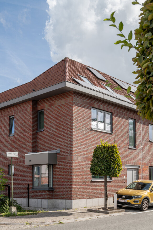 Hoekwoning van de sociale woningen in Wetteren, in rode baksteen met grijze ramen, een kleine luifel boven de deur en zonnepanelen op het pannendak