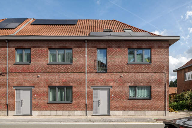 Sociale woningen in Wetteren onder één pannendak, met gevels in rode baksteen, grijze voordeuren en ramen, en zonnepanelen op het dak