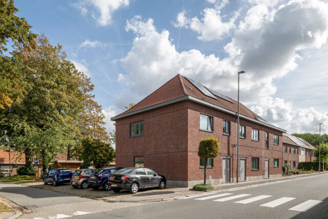 Hoekgebouw met sociale woningen in Wetteren, in rode baksteen met een pannendak, aan een kruispunt met bomen, geparkeerde auto's en een zebrapad