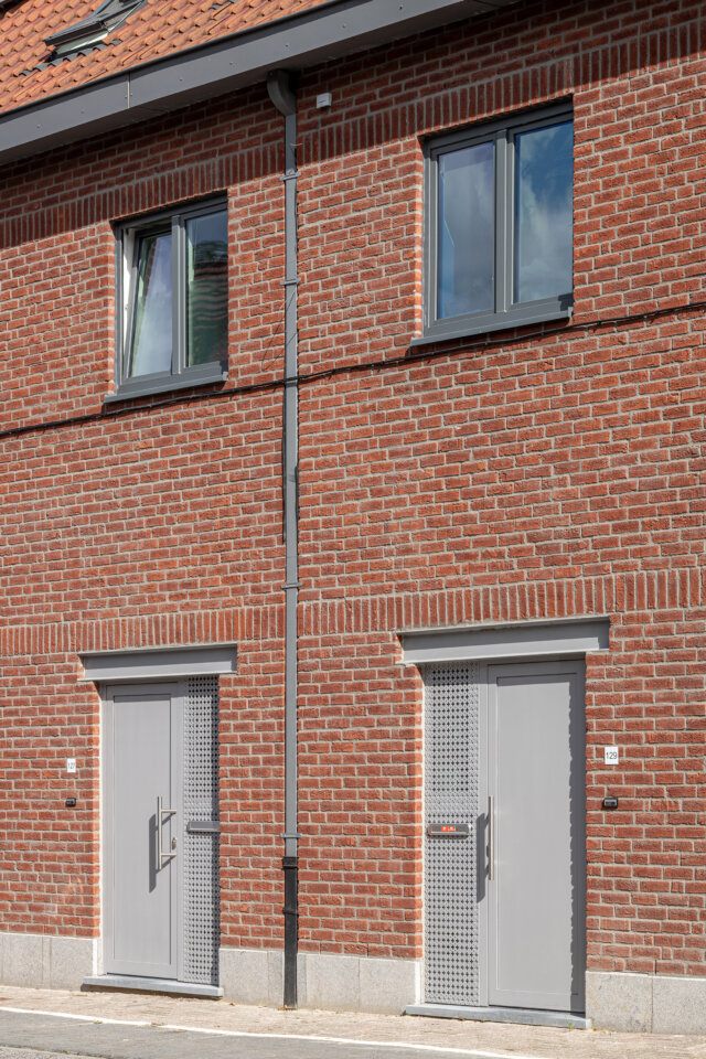 Twee grijze voordeuren naast elkaar in de rode bakstenen gevel van twee gekoppelde sociale woningen in Wetteren, met grijze ramen op de verdieping