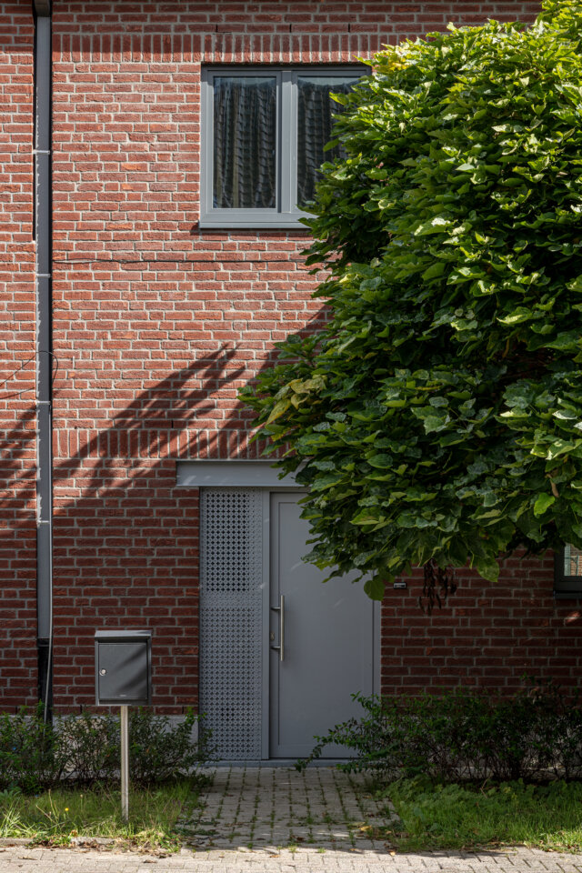 Grijze deur in de rode bakstenen gevel van een sociale woning in Wetteren, met een laag hekje, een brievenbus en een boom die schaduw werpt
