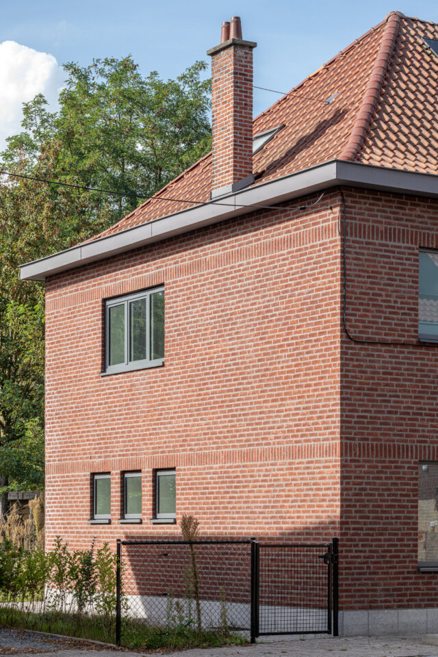 Gerenoveerde sociale woning in Wetteren in rode baksteen, met een schoorsteen, nieuwe grijze ramen, een pannendak en een zwart hekje tussen het groen