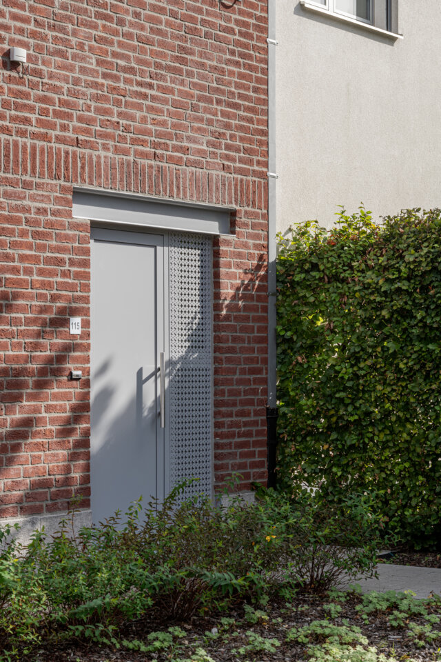 Grijze voordeur in de rode bakstenen gevel van een sociale woning in Wetteren, met een haag en beplanting ernaast