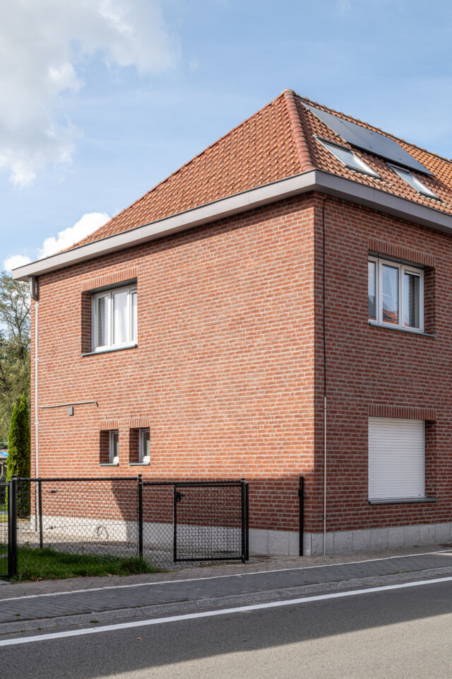 Zijgevel van de sociale woningen in Wetteren, in rode baksteen met witte ramen, een pannendak met zonnepanelen en een zwart hekwerk langs de straat