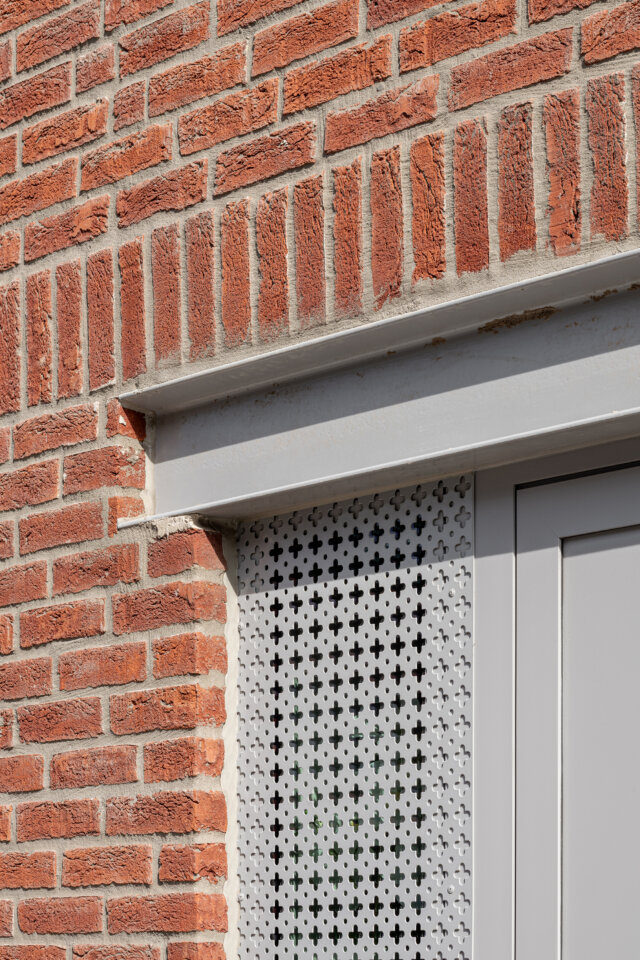 Detail van een raam met grijs kader en een geperforeerd metalen luik in de rode bakstenen gevel van de sociale woningen in Wetteren